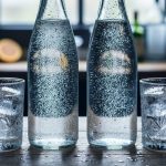Two chilled carbonated water bottles and glasses with condensation and rising bubbles on a bar countertop under soft daylight.
