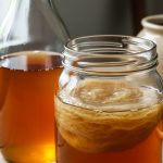 Close-up of kombucha in a glass jar showing a SCOBY floating in amber sweet tea with homebrewing tools blurred in the background.