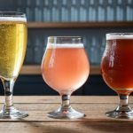 Three sour beer glasses in different colors with visible carbonation and condensation on a wooden tasting table, photographed in a dim taproom setting.